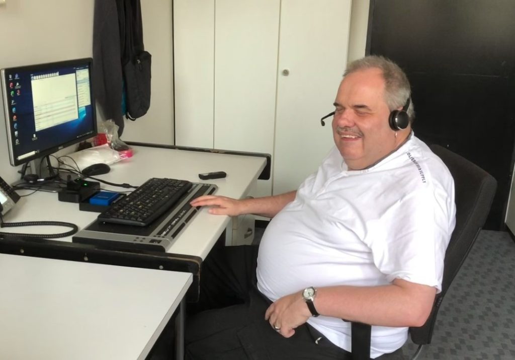 Ein Mann, es ist Bernd Hermandung, mit grauen, kurzen Haaren und einem Oberlippenbart. Er trägt ein weißes T-Shirt und sitzt in einem Stuhl vor einem Computer mit einer Tastatur für Blinde. Dabei hat er ein schwarzes Headset auf.