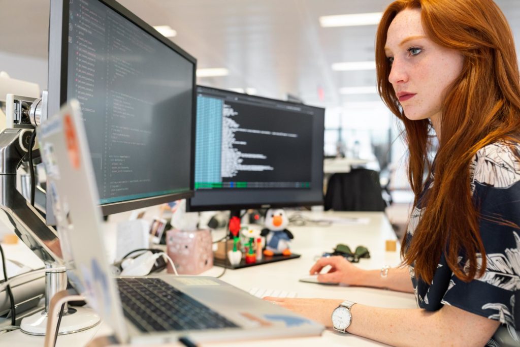 Frau mit langen, roten Haaren und dunklem T-Shirt mit Blumenmuster sitzt am Arbeitsplatz mit mehreren Monitoren.