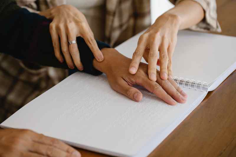Ein Buch mit Blindenschrift liegt aufgeklappt auf einem Tisch. Zwei Hände führen eine andere Hand über die Schriftzeichen.