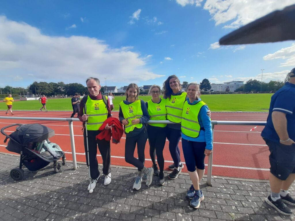 Die Gruppe steht vor dem Geländer der Laufbahn, trägt die gelben BFW Düren-Westen und lächelt in die Kamera.