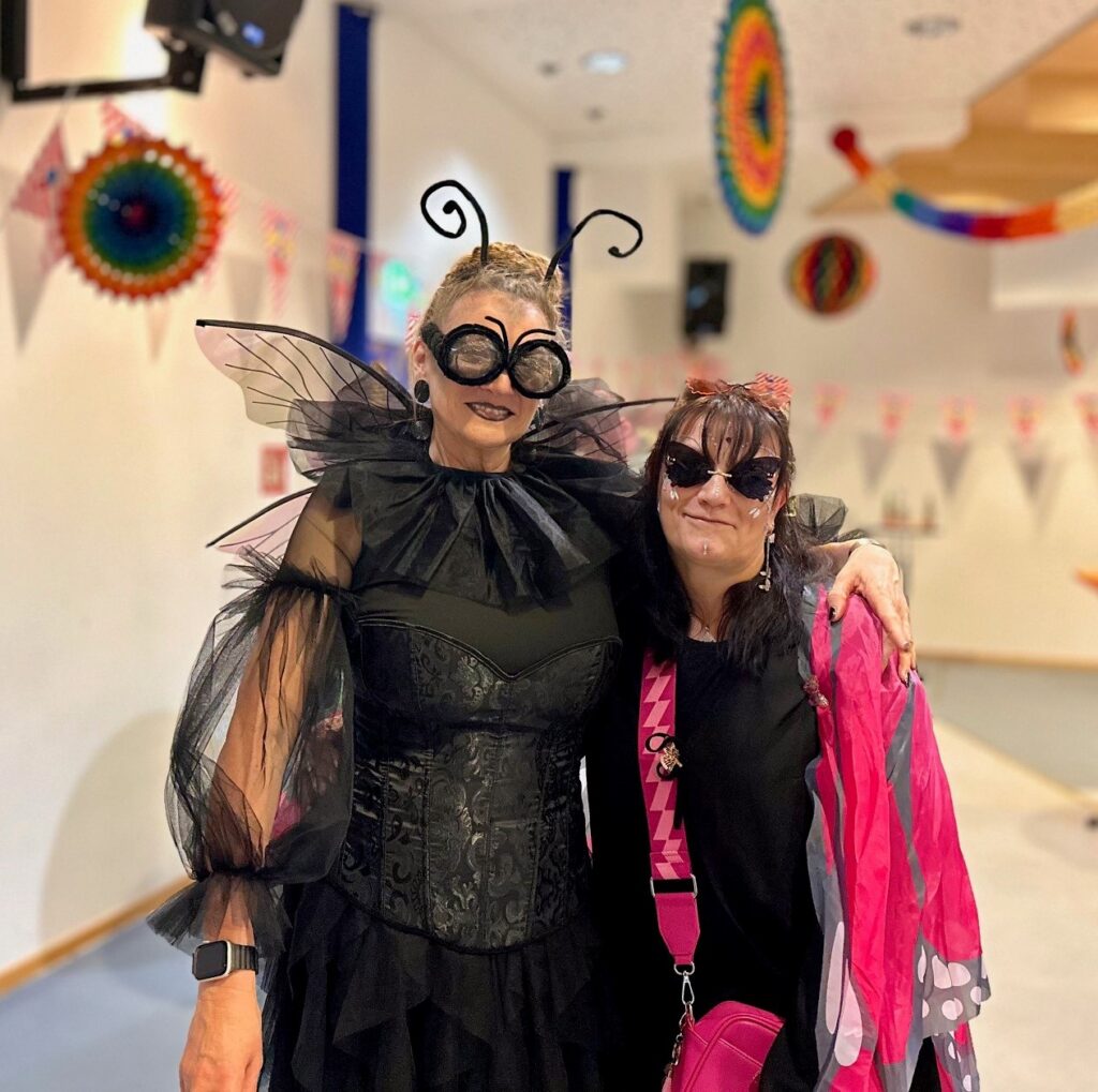 Zwei Frauen stehen lächelnd nebeneinander. Eine trägt ein schwarzes Insektenkostüm mit Flügeln und großen Brillengläsern, die andere ein dunkles Outfit mit pinkem Schal und Sonnenbrille. Der Raum ist bunt geschmückt.