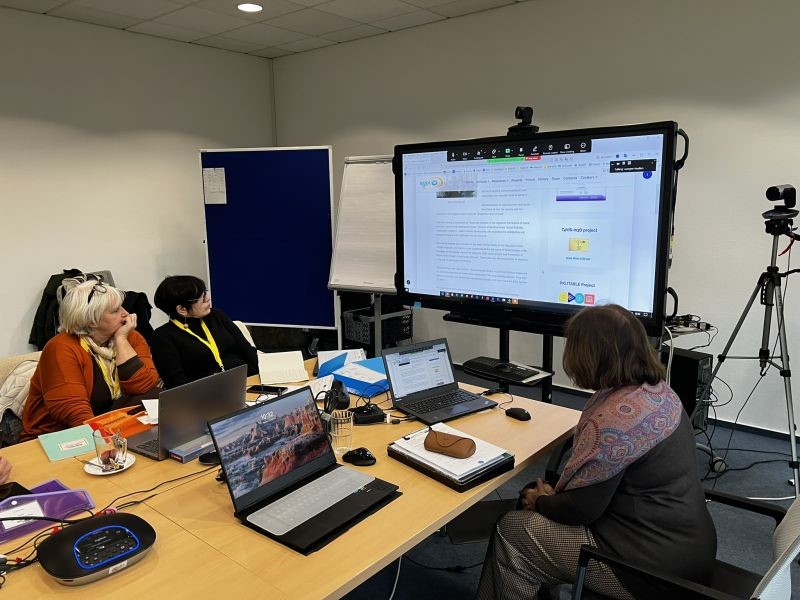 Mehrere Personen sitzen an einem Besprechungstisch mit Laptops und schauen auf einen großen Bildschirm mit einer Präsentation während eines Workshops.