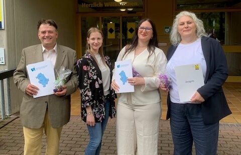Drei Absolventinnen und Absolventen der Umschulung zum Verwaltungsfachangestellten gemeinsam mit einer Ausbilderin vor dem Eingangsbereich des BFW Düren. Einige halten ihre Abschlussunterlagen in den Händen. Die Gruppe lächelt in die Kamera.