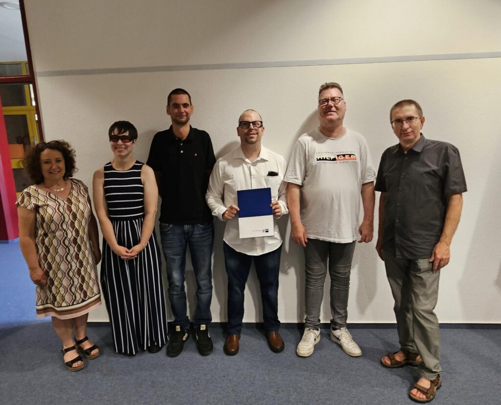 Gruppenfoto von fünf Absolventinnen und Absolventen gemeinsam mit einer Ausbilderin. Die Gruppe steht vor einer hellen Innenwand im BFW Düren. Eine Person hält eine blaue Abschlussmappe in den Händen. Die Teilnehmenden blicken lächelnd in die Kamera.