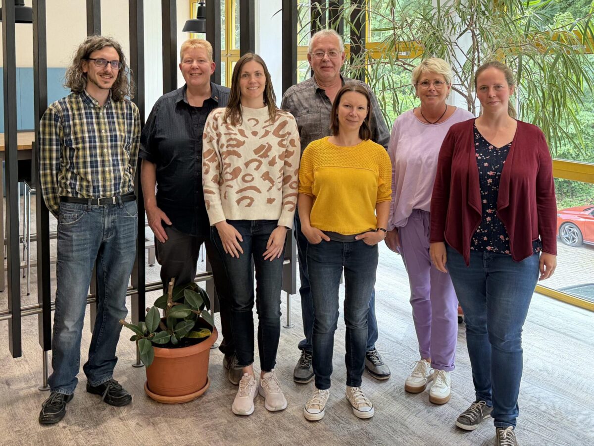 Gruppenfoto der Mitglieder des Betriebsrats des BFW Düren in einem hellen Innenbereich mit großen Fenstern und Pflanzen. Sieben Personen stehen nebeneinander und blicken in die Kamera. Im Vordergrund befindet sich eine Topfpflanze.