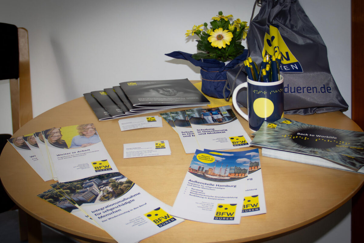 Auf einem runden Tisch liegen verschiedene Informationsbroschüren, Flyer und Visitenkarten des BFW Düren. Daneben stehen Werbeartikel wie eine Tasse mit Stiften, eine Tragetasche mit BFW-Logo sowie eine Topfpflanze mit gelben Blüten. Die Materialien sind für einen Informationstag am Standort Hamburg vorbereitet.