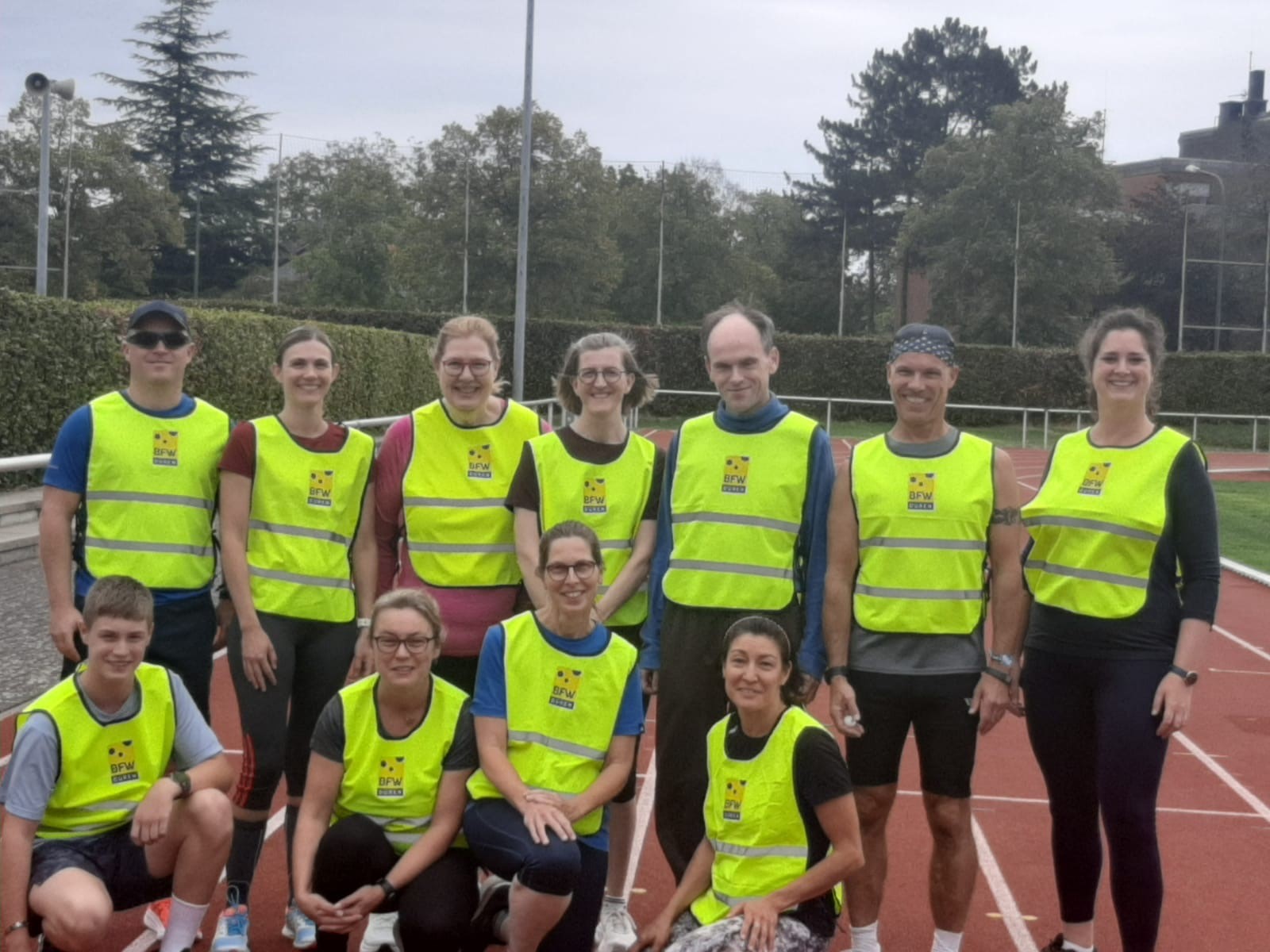 Gruppenfoto von Mitarbeitenden des BFW Düren beim Inklusionslauf Düren. Die Teilnehmenden stehen und knien gemeinsam auf einer Laufbahn und tragen leuchtend gelbe Warnwesten mit BFW-Logo. Insgesamt waren 17 Kolleginnen, Kollegen und Angehörige für das BFW beim Inklusionslauf dabei.