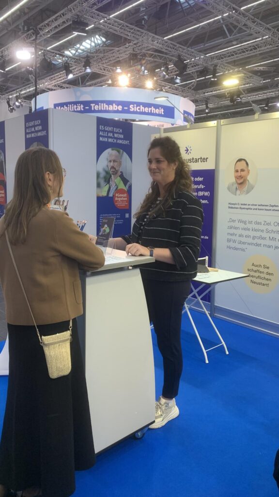 Zwei Frauen im Gespräch an einem Messestand auf der REHACARE. Im Hintergrund sind Informationsmaterialien und Messewände zum beruflichen Neustart und zum Thema Gewaltprävention zu sehen. Über dem Stand steht „Mobilität – Teilhabe – Sicherheit“.
