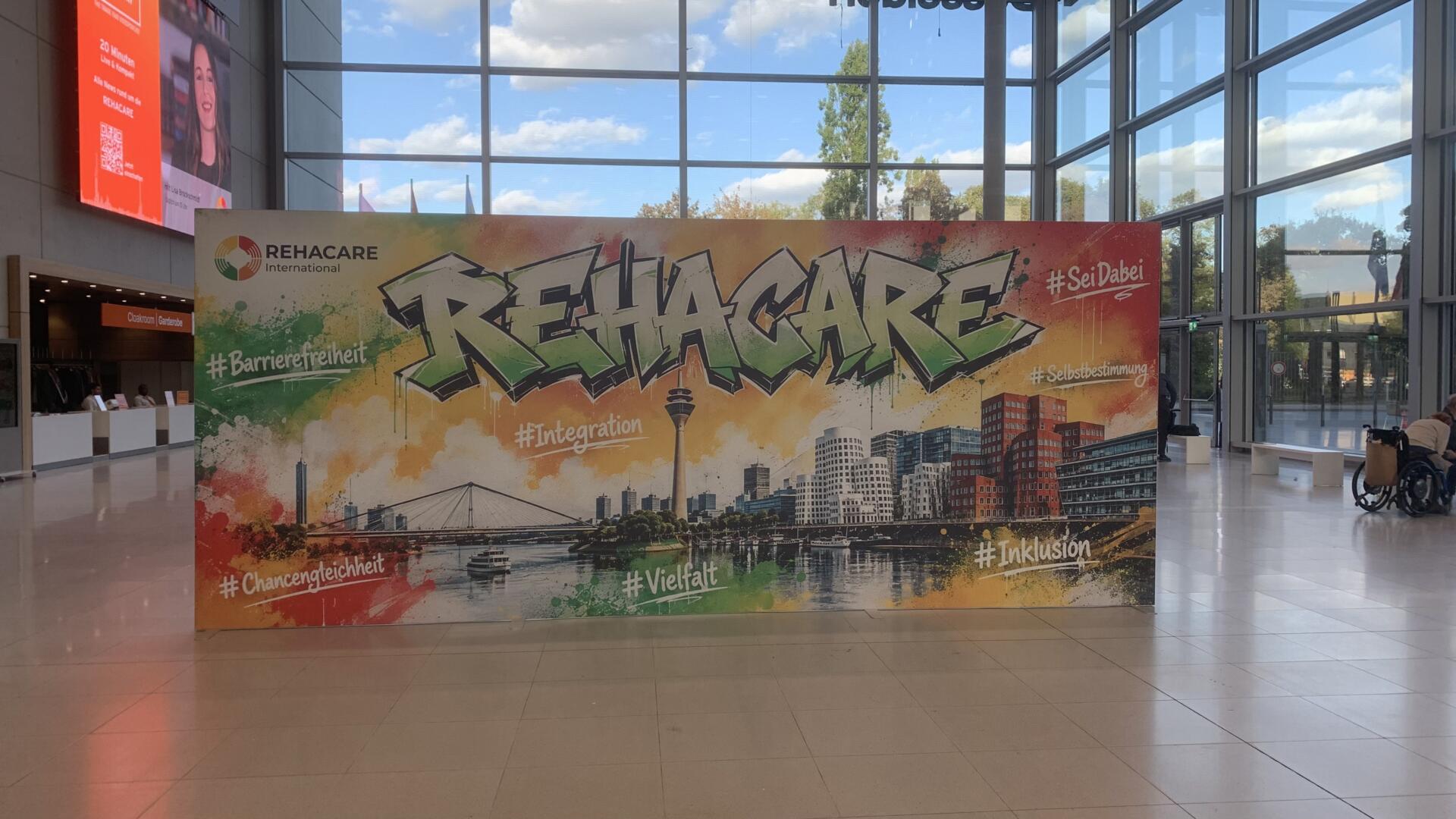 Große, farbenfrohe REHACARE-Fotowand in einer Messehalle mit einer illustrierten Skyline von Düsseldorf. Rund um den Schriftzug „REHACARE“ stehen Begriffe wie Barrierefreiheit, Integration, Chancengleichheit, Vielfalt, Inklusion und Selbstbestimmung.